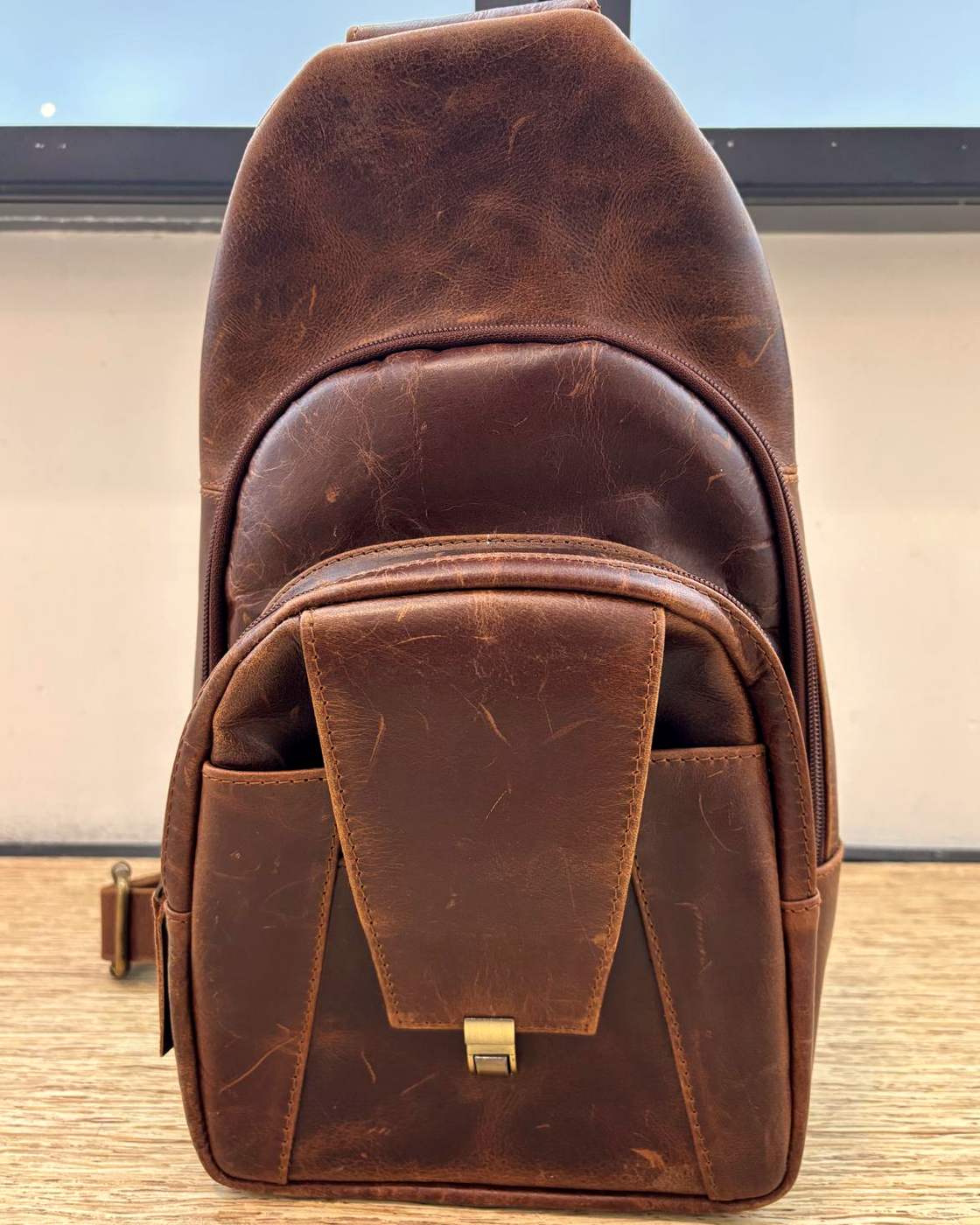 Real photo of the Courier Sling standing upright on a wooden table, front on, showing the teardrop shape, the curved main zip running around the front, the flap pocket at the bottom with its metal clasp, and slot pockets on either side of the flap.
