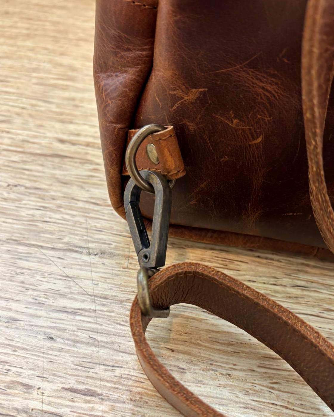 Close-up real photo of the Courier Sling strap hardware: a swivel snap hook clipped through a ring on a stitched leather tab at the bottom corner of the bag, with the strap running away across the table.