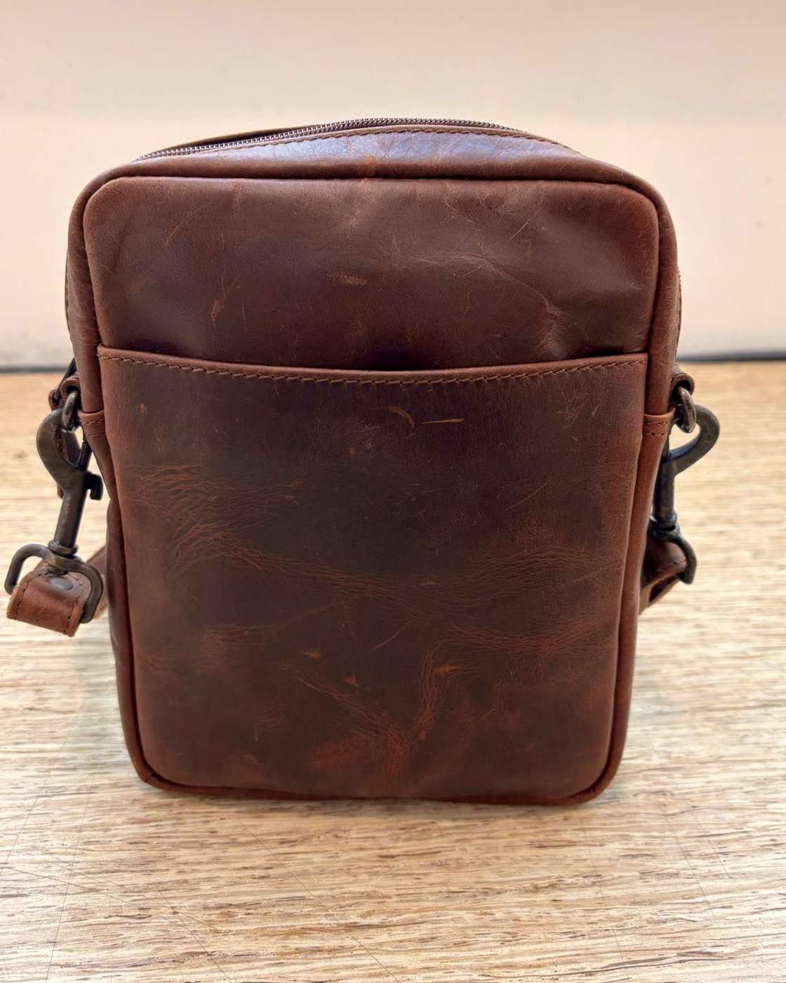 Real photo of the YYZ Satchel standing on a wooden table, showing the face with an open slip pocket running the full width under the top panel, with strap clips at both upper side seams.