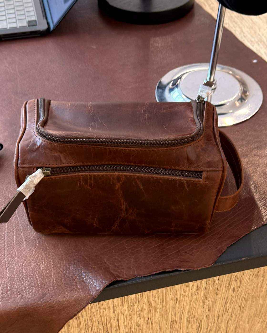 Real photo of the brown leather dopp kit on a leather hide on the shop counter, three-quarter view, showing the top zip, the front zip pocket and the leather zip pull with its shop tag still on.