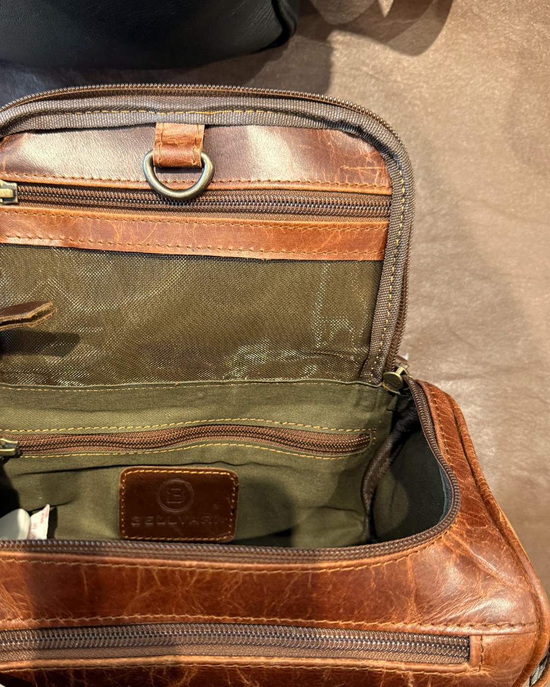 Real photo of the dopp kit opened, lid up, showing the olive lining, the mesh zip pocket inside the lid, the brass D-ring, the zip pocket on the back wall and the Bellvari leather patch.