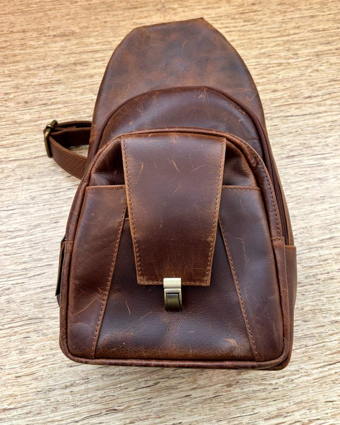 Real photo of the Courier Sling lying flat on a wooden table, shot from above, showing the whole bag front face up with the flap pocket and clasp, the slot pockets beside it, and the strap running off to one side.