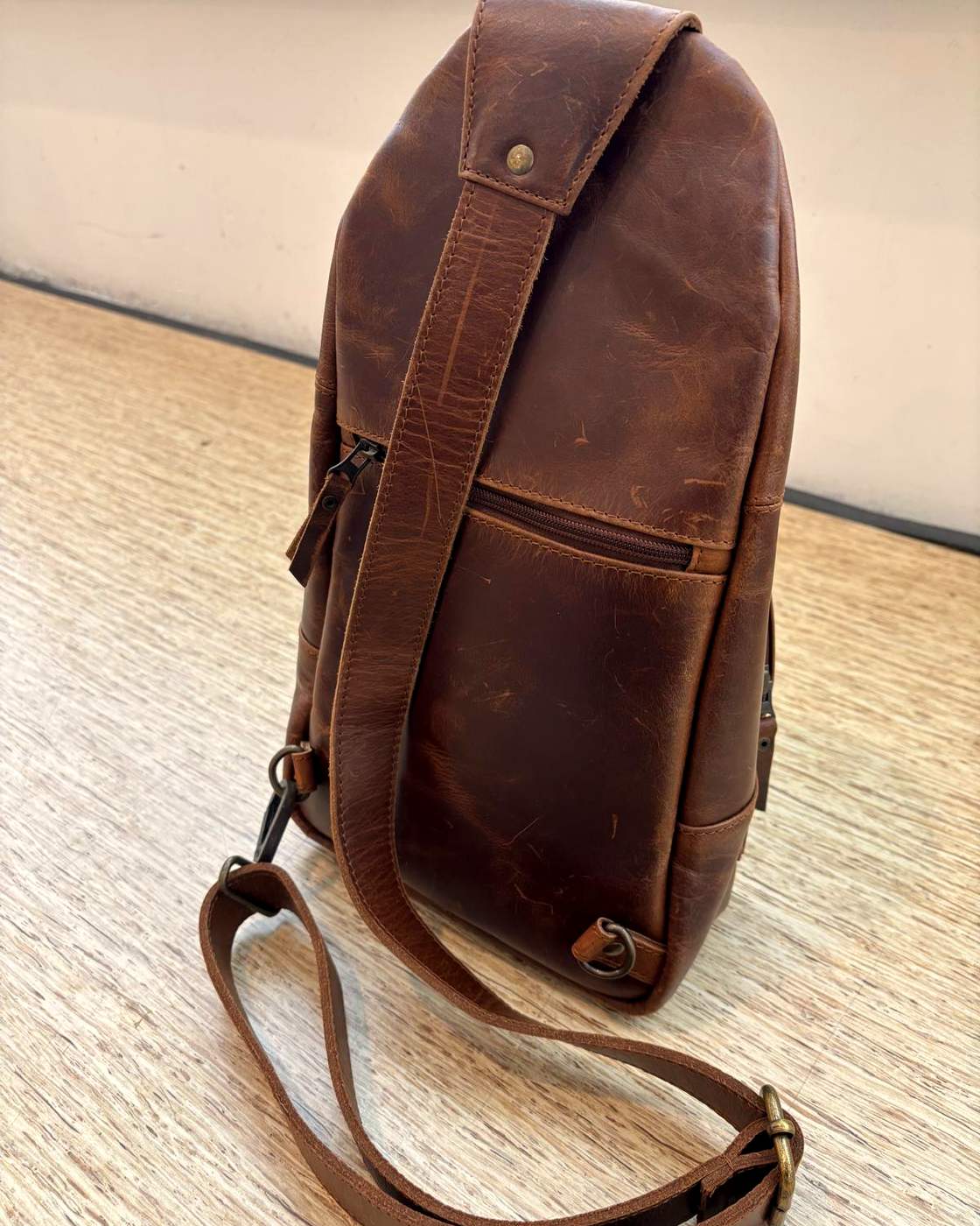 Real photo of the Courier Sling standing on a wooden table, seen at an angle from behind, showing the depth of the bag, the wide strap panel running down the back, the zipped back pocket and the loose strap laid out on the table.