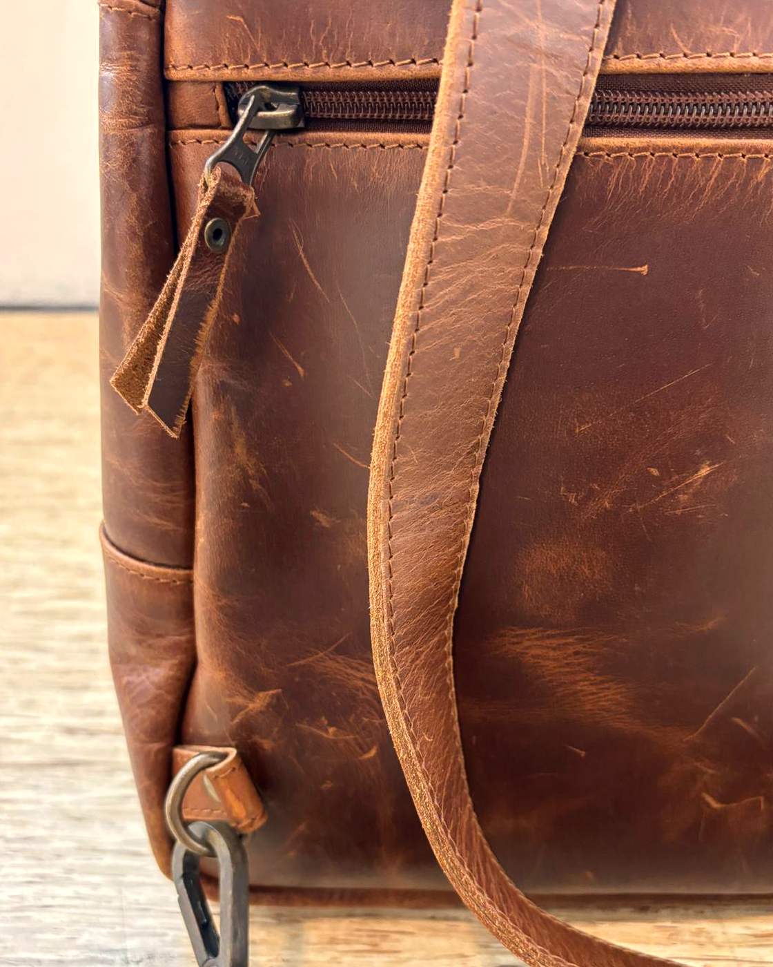 Close-up real photo of the zipped pocket on the back of the Courier Sling, showing the zip running across the panel, its metal slider and leather pull tab, and the strap passing over it.