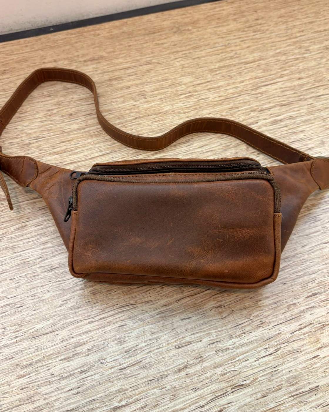 Real photo of the Nomad Sling lying flat on a pale wooden table with its strap laid out in a loop behind it, showing the crescent-shaped body, the curved-top front pocket and the full length of the strap.