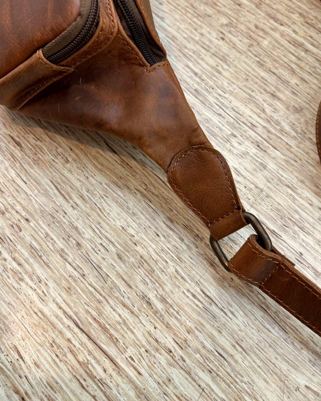 Close-up real photo of the end of the Nomad Sling, where the body tapers into a stitched leather tab holding a metal ring, with the strap threaded through it.